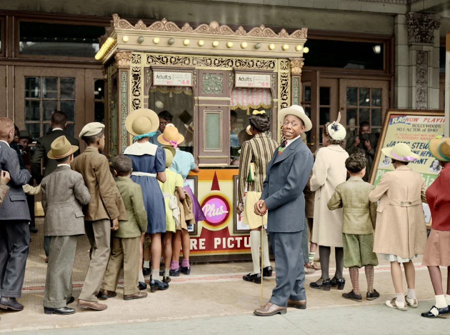 A group of children and adults, dressed in vintage clothing, gather at a theater box office. One man in a suit and hat stands looking toward the camera, while others face the booth, waiting to buy tickets.