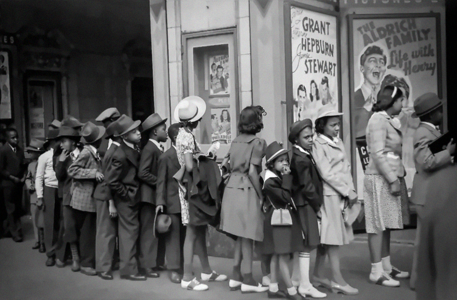 A group of children in coats and hats stand in line outside a theater, waiting to enter. Movie posters with classic film stars are visible on the wall behind them.