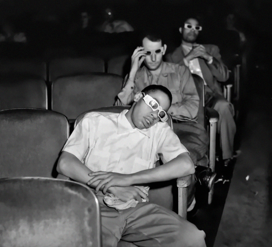 Three men wearing 3D glasses sit in a mostly empty theater; one man in front is asleep with arms crossed, while the others adjust their glasses and look forward. The image is in black and white.