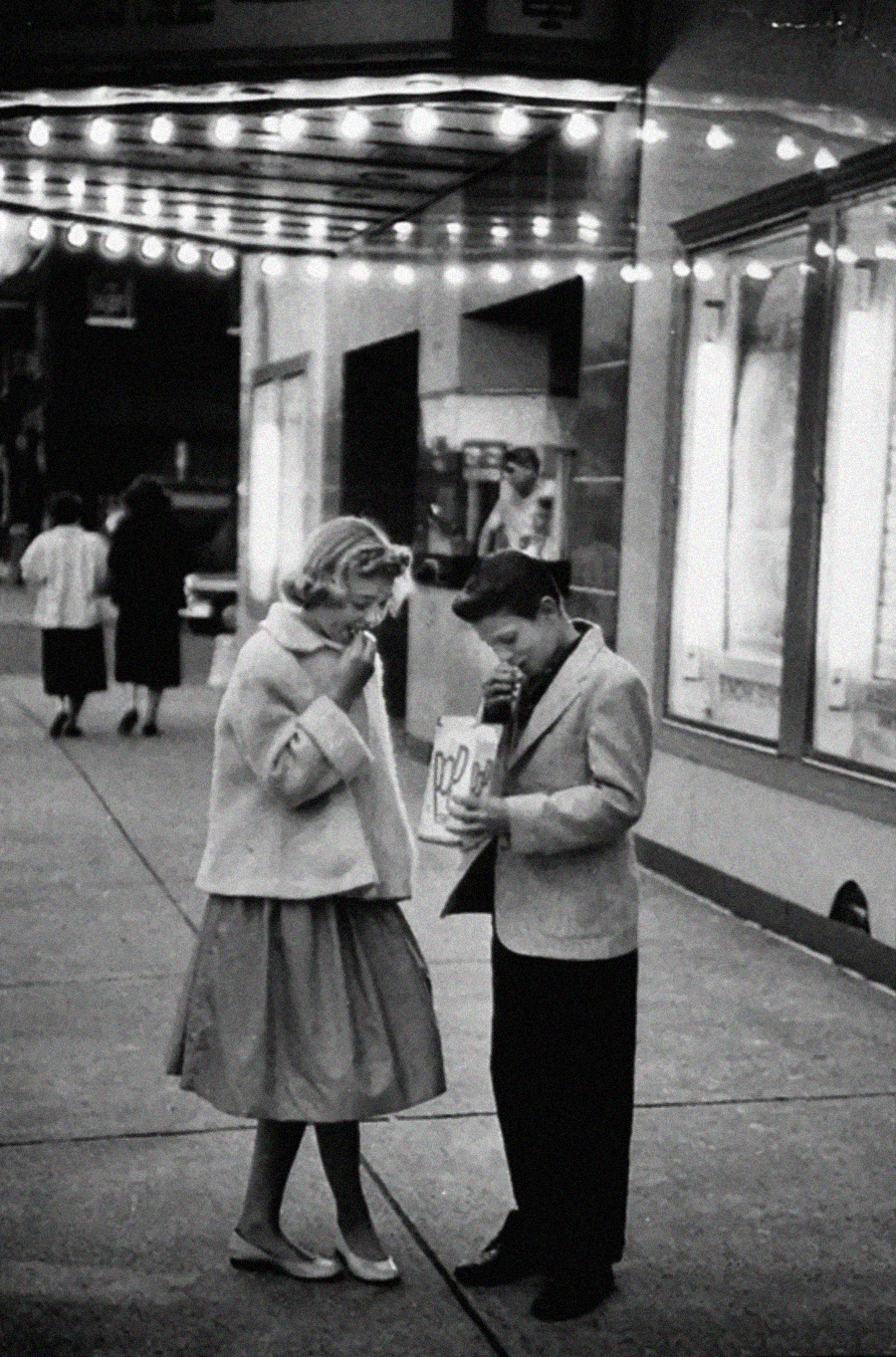 A young boy and girl in vintage clothing stand outside a lit-up theater at night, sharing popcorn from a bag. Other people walk by in the background.
