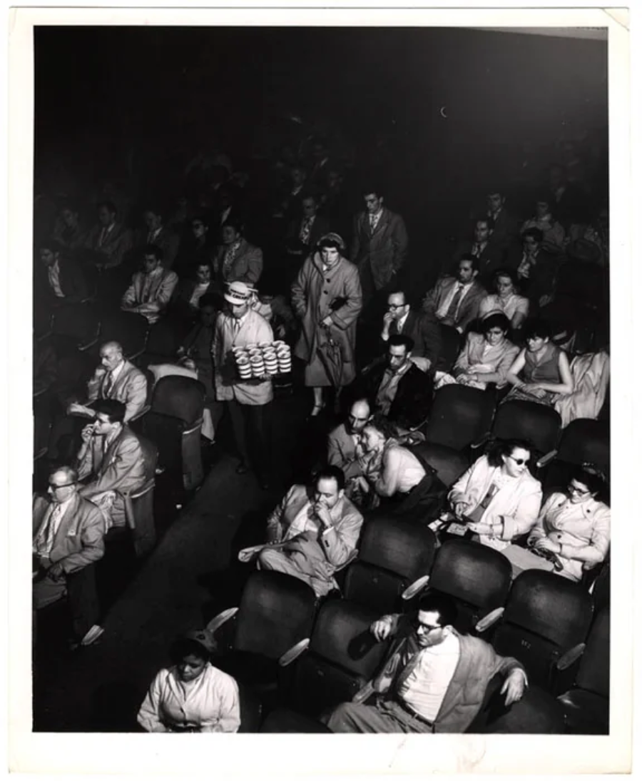 Black-and-white photo of a theater audience wearing 3D glasses. A person walks down the aisle carrying a tray of drinks and snacks. Most people are looking toward the front, seated in rows of chairs.