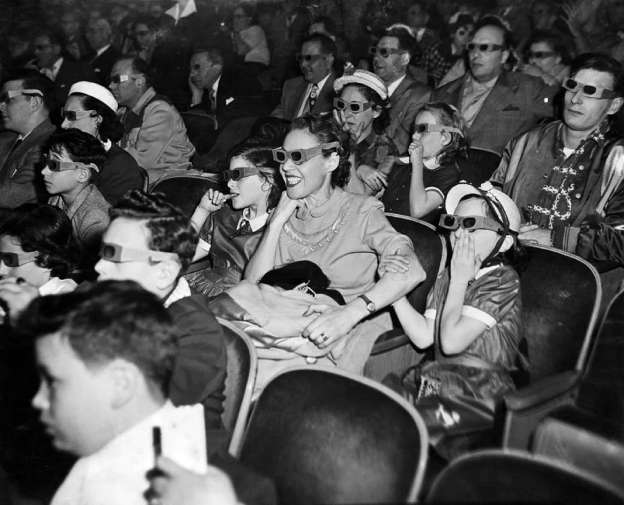A large audience, including adults and children, sits in rows of a theater wearing 3D glasses, watching a movie. The crowd appears engaged and some children look surprised or excited.