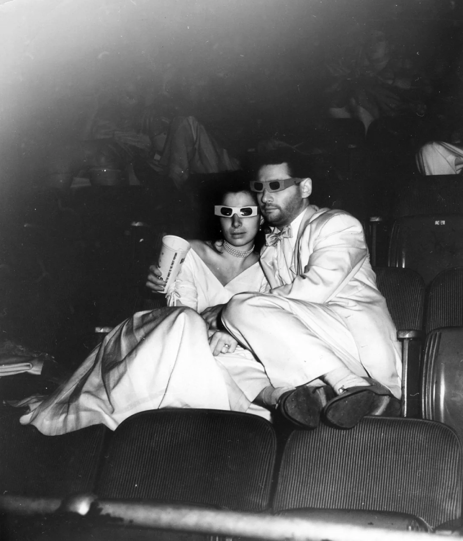 A couple dressed in formal clothing sits closely together in theater seats, wearing 3D glasses and holding a drink, watching a movie in a dark cinema.