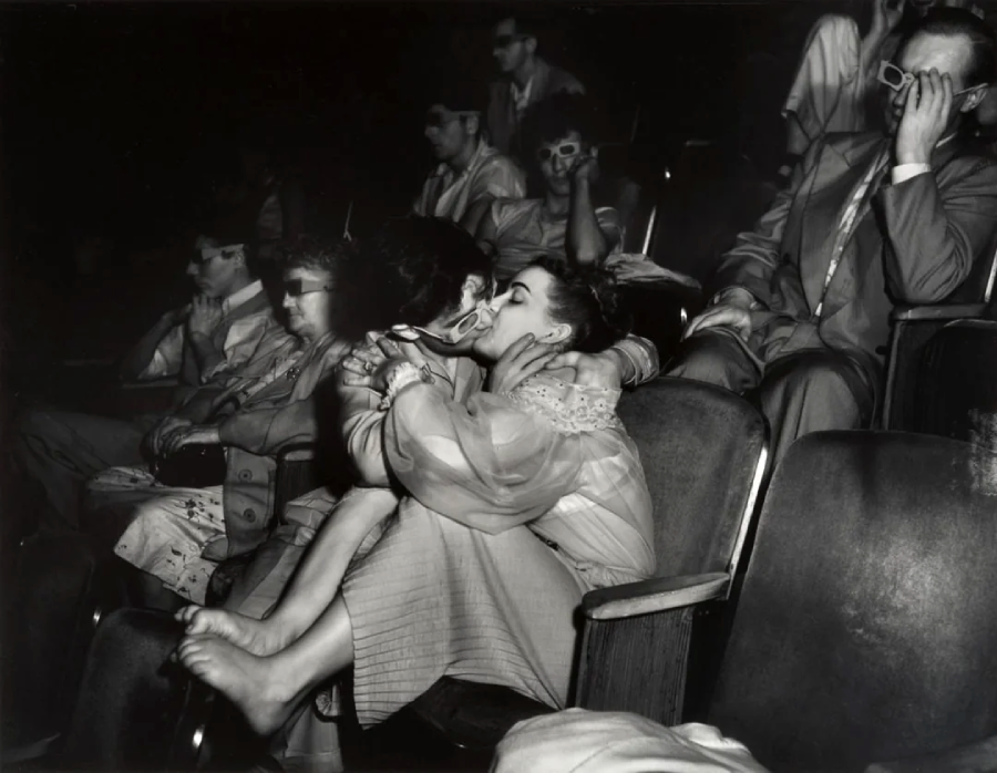 A black-and-white photo shows a young couple kissing passionately in a movie theater, with the woman sitting barefoot on the man's lap. People around them, wearing 3D glasses, are focused on the screen.