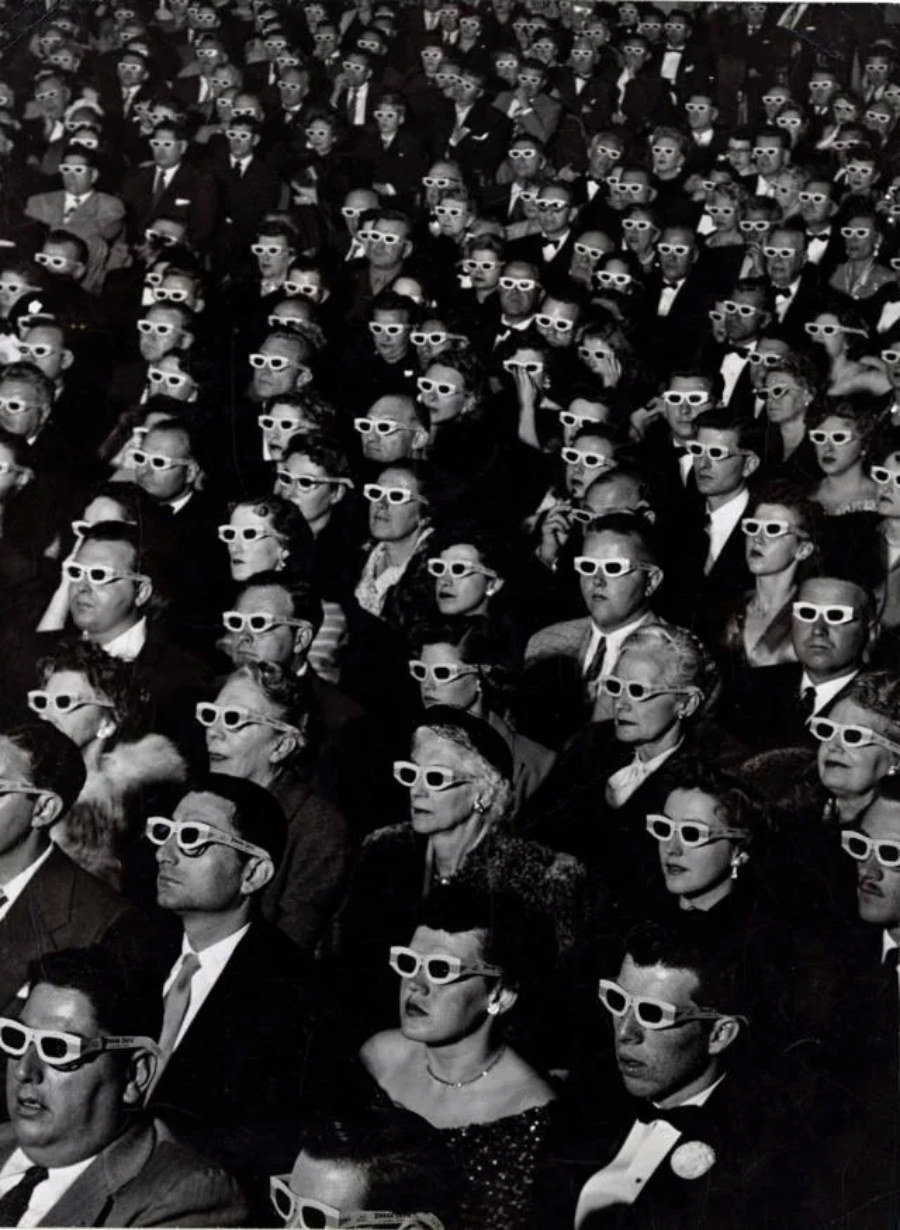 A large crowd of people sits in a theater, all wearing identical 3D glasses and looking toward the screen. The image is in black and white and the audience appears focused and attentive.
