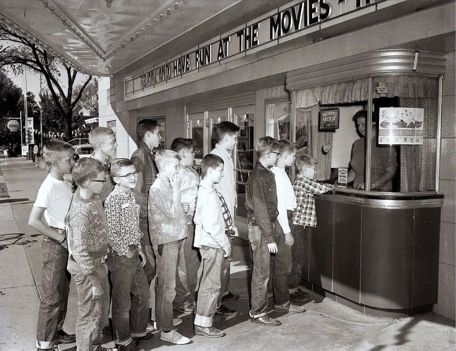 A group of boys in vintage clothing stand in line at a movie theater box office, waiting to buy tickets from a cashier behind a window. The scene appears to be from the 1950s or 1960s.