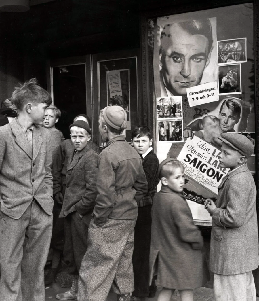 A group of boys in old-fashioned clothing stand outside a building with movie posters, including one featuring a man's face and the title "SAIGON." The boys appear to be waiting, some looking at the camera.