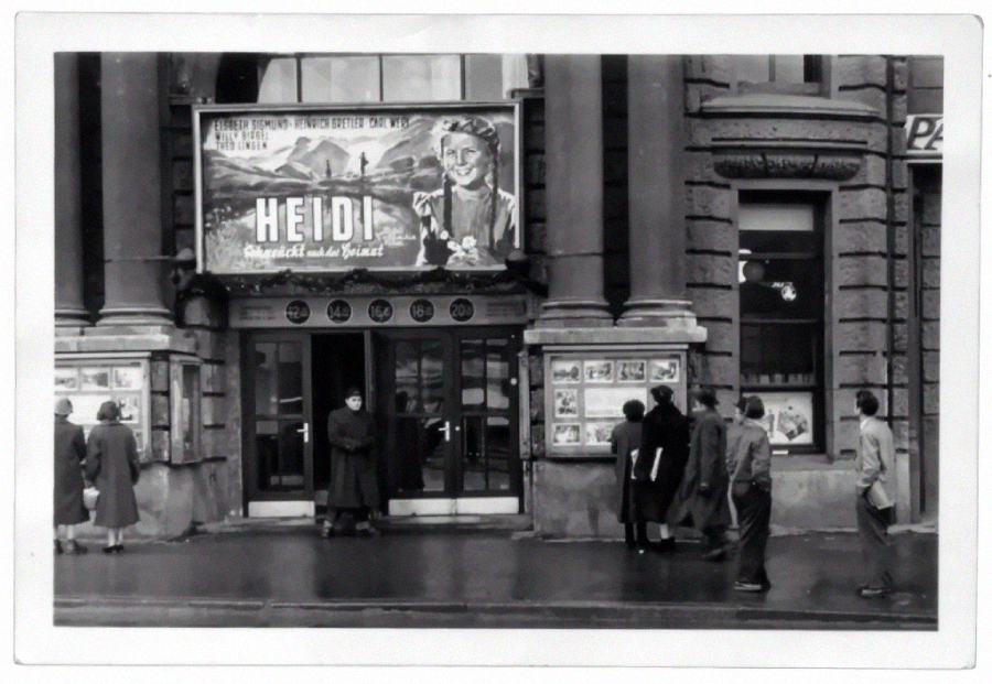 Black-and-white photo of people standing outside a theater entrance, looking at posters. Above the entrance is a large “Heidi” movie poster featuring a girl and alpine scenery. The street and building appear old-fashioned.