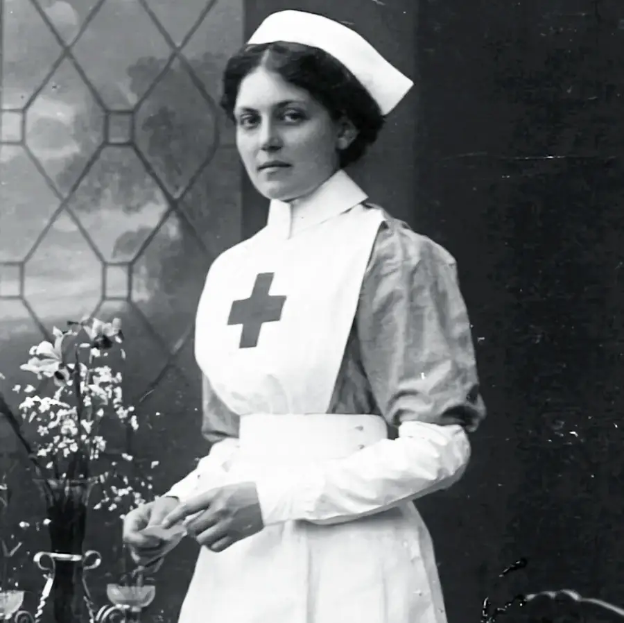 A black-and-white photo of a nurse in an old-fashioned uniform with a Red Cross emblem, standing indoors near a table with flowers and a stained glass window in the background.