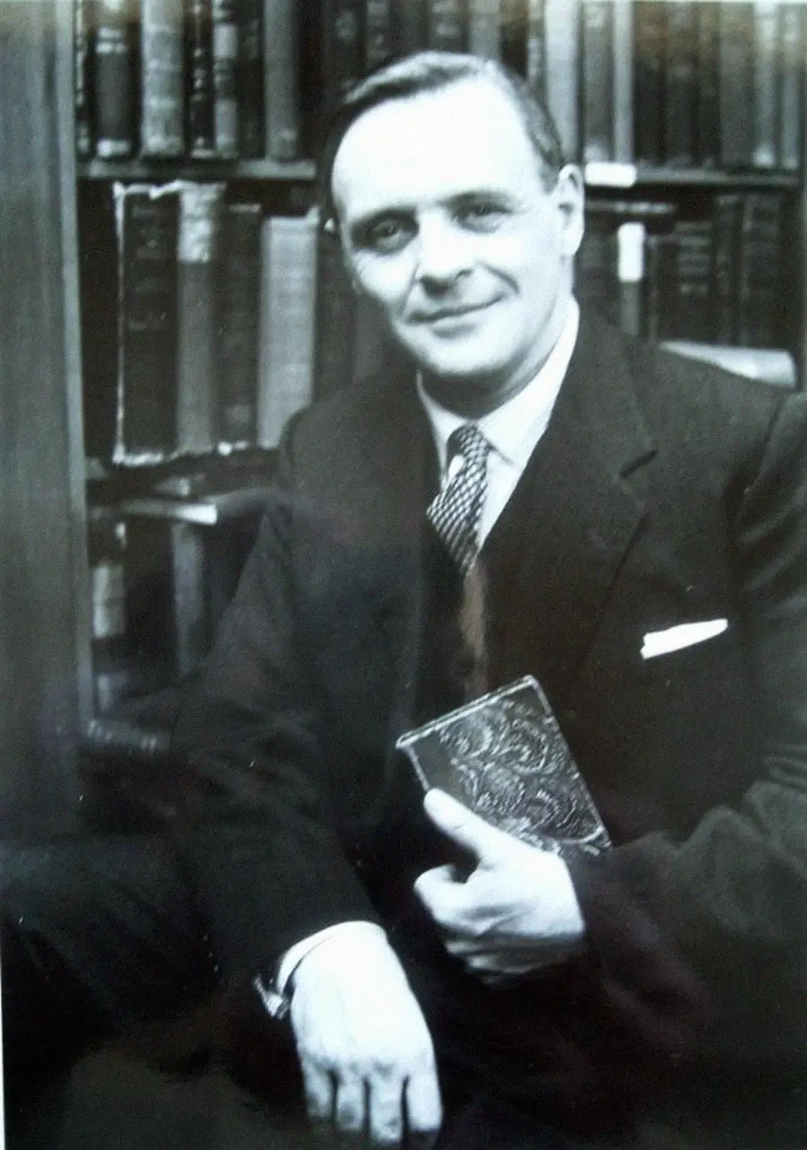 A man in a suit sits in front of a bookshelf, smiling and holding a book with an ornate cover in his hand. The photo is in black and white.