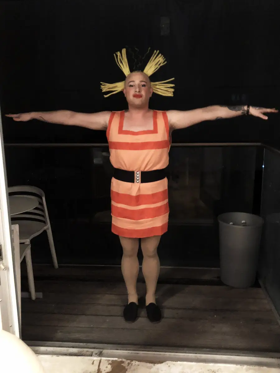 A person stands with arms outstretched, wearing a striped orange dress, beige tights, black shoes, and a headpiece resembling yellow hair spikes. The background includes stacked chairs and a trash can on a porch.