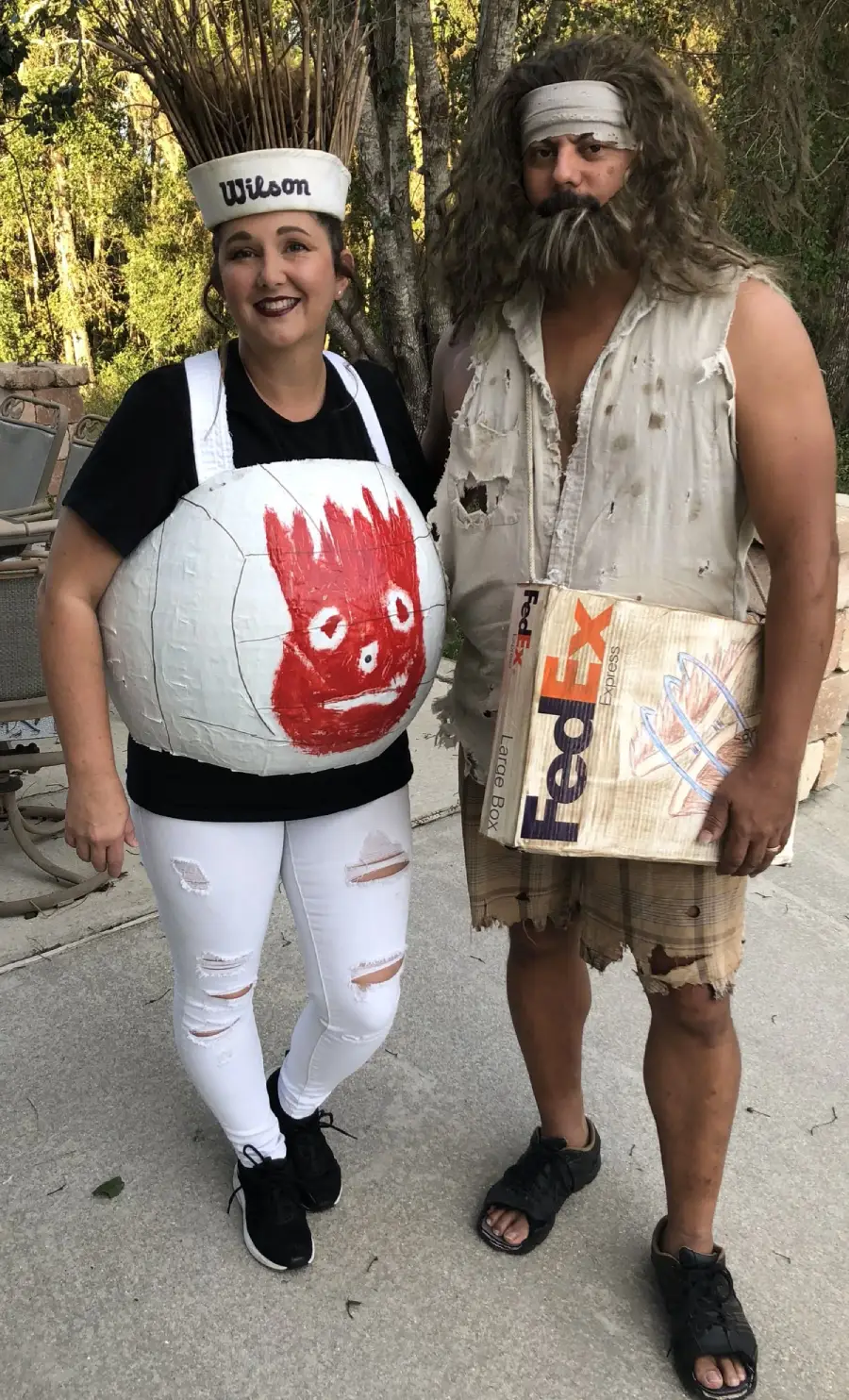 Two people in costumes: one dressed as a volleyball with a red face labeled "Wilson," the other as a bearded castaway in tattered clothes holding a FedEx box, referencing the movie "Cast Away." They are standing outdoors.