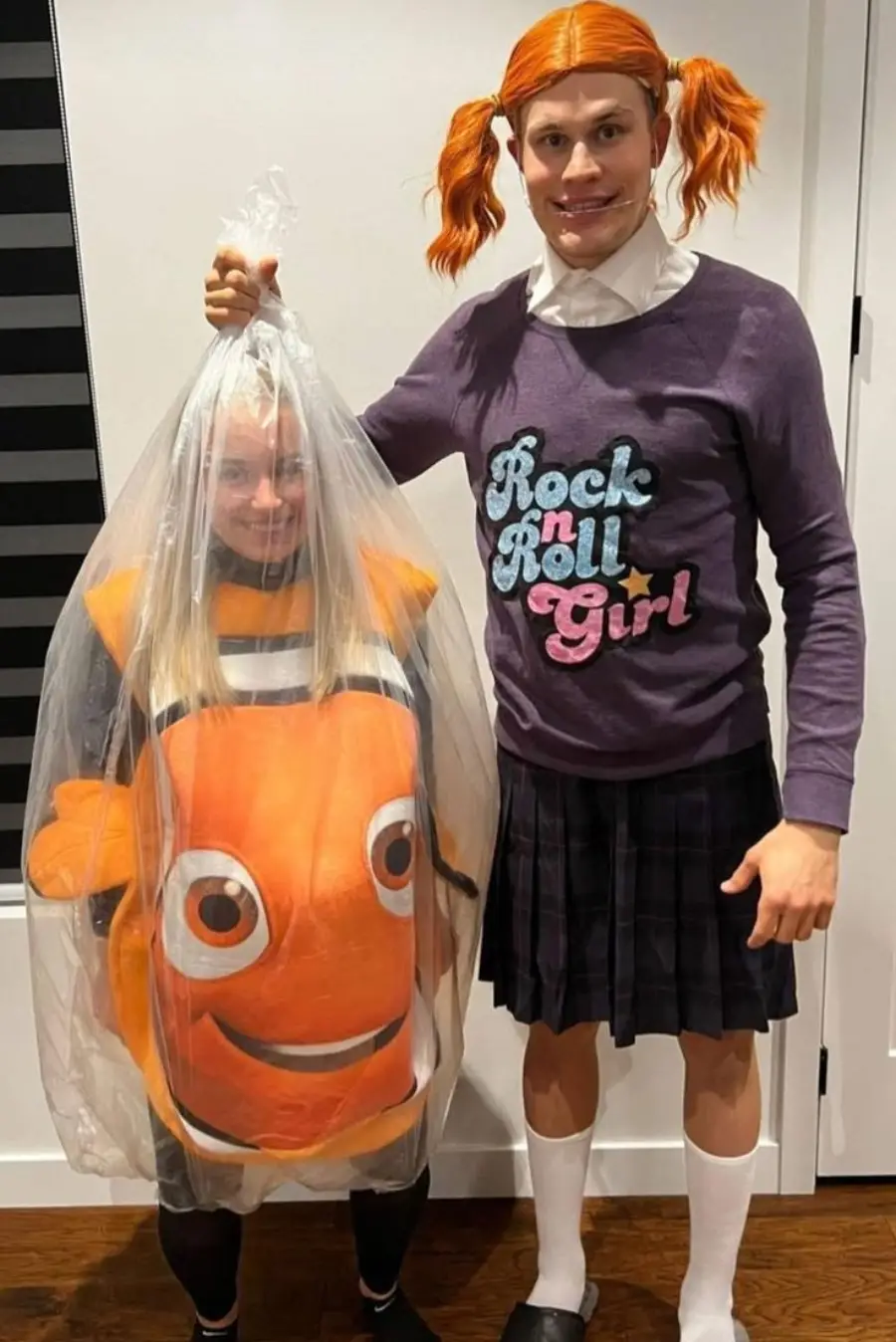 Two people in costumes: one dressed as Nemo from "Finding Nemo" inside a clear plastic bag, the other in a purple “Rock n Roll Girl” shirt, black skirt, and orange pigtail wig, both posing and smiling indoors.