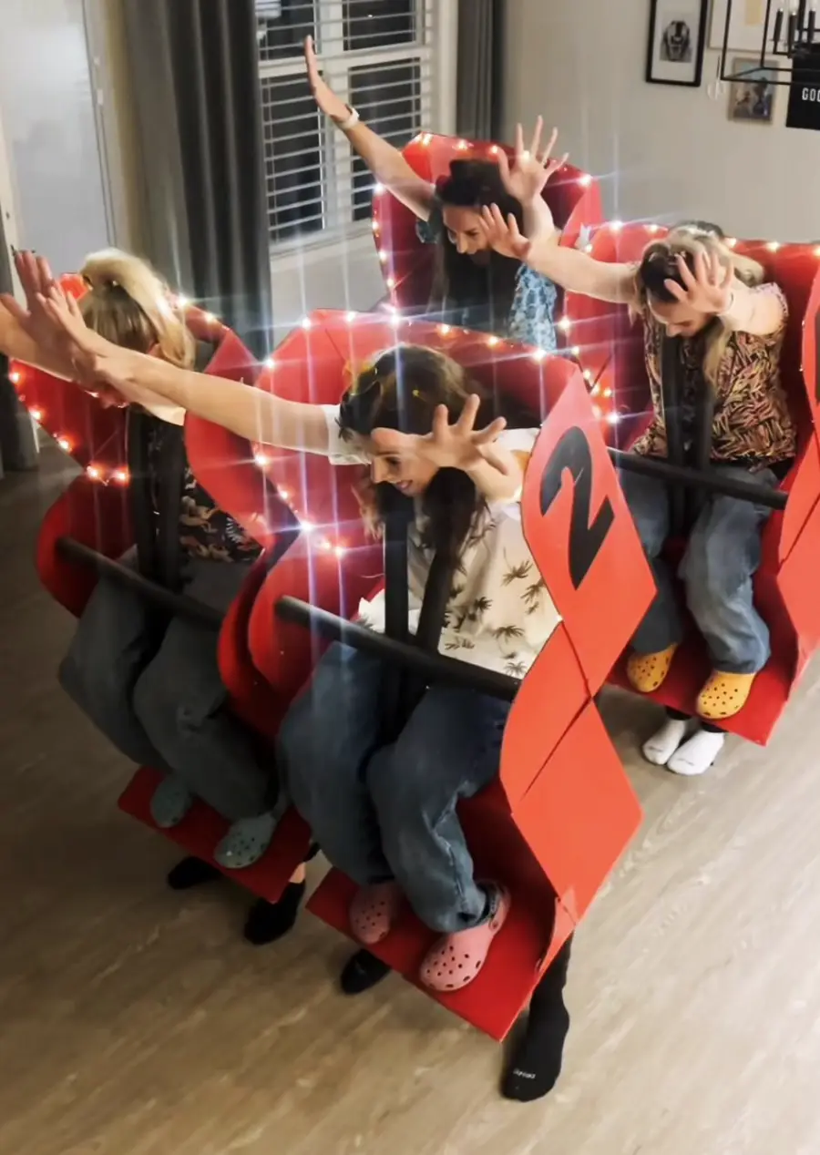 Four people sit in homemade red cardboard roller coaster seats, wearing seatbelts and raising their hands as if on a ride. String lights decorate the setup, and two seats have the number 2 on them. They appear to be indoors.