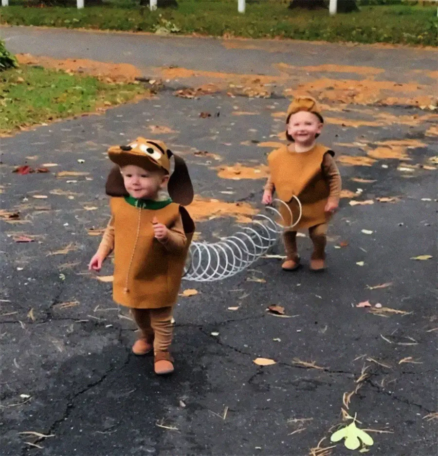 Two young children wearing matching Slinky Dog costumes walk outside on a driveway, connected by a stretchy silver coil. Both are smiling and dressed in brown outfits with dog ears and tails. Leaves are scattered on the ground.