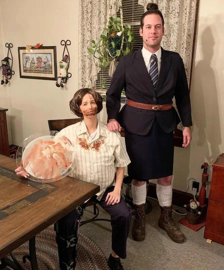 Two people in a home dressed in costumes: one seated with a fake mustache, messy shirt, and a plate of bacon; the other standing, wearing a suit jacket, skirt, tie, and boots. The scene has quirky, playful energy.