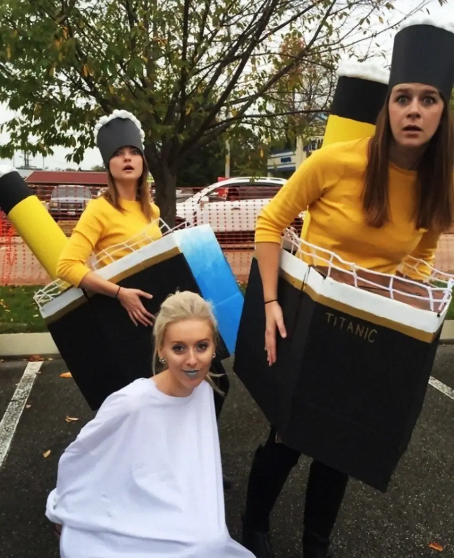 Three women in a parking lot wear creative Halloween costumes. Two dress as the Titanic ship with yellow smokestacks, while the third, dressed in white, poses as an iceberg. Trees and cars are visible in the background.