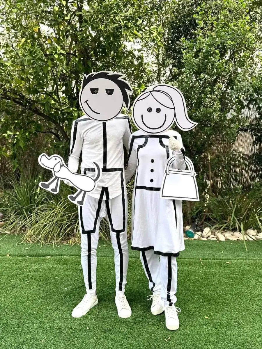 Two people dressed as cartoon characters with black-and-white outfits and large, simple cartoon face masks. One holds a cartoon dog cutout, and the other holds a cartoon handbag. They stand on grass with trees in the background.