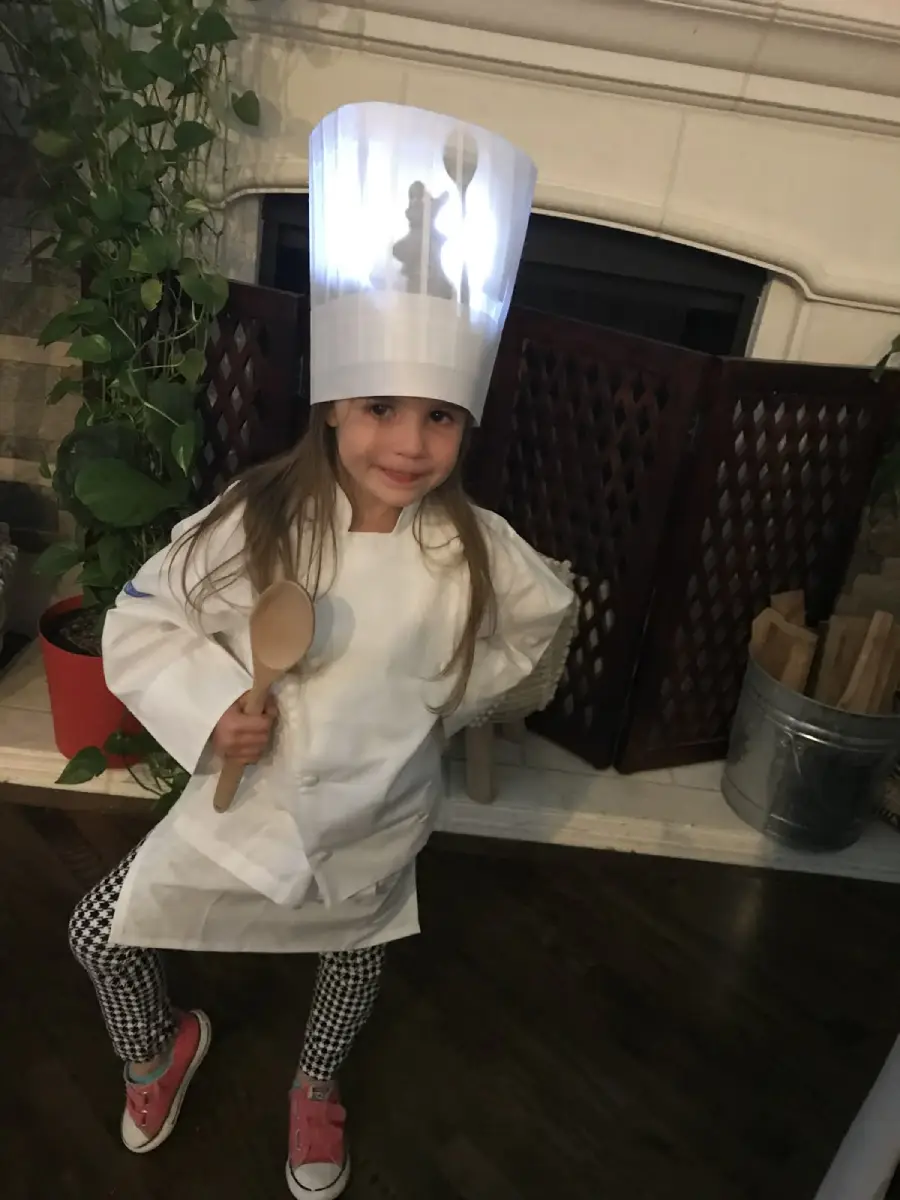 A young girl dressed as a chef poses indoors, wearing a tall paper chef hat and white chef coat, holding a wooden spoon, and smiling. She stands in front of a fireplace with plants and a metal bucket nearby.