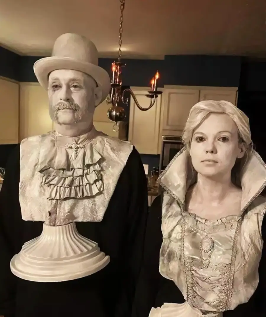 Two people dressed as vintage white marble busts, with detailed period costumes and makeup to appear like statues. They stand side by side in a kitchen, with a chandelier and cabinets in the background.