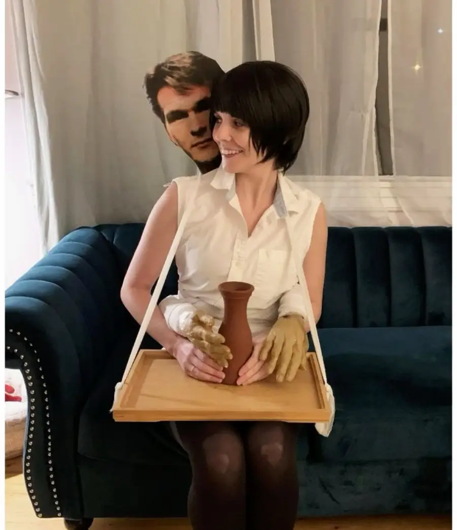 A woman on a blue couch wears a costume resembling a famous pottery scene, with a cardboard cutout of a man behind her, fake arms around her, and holding a clay vase on a tray.
