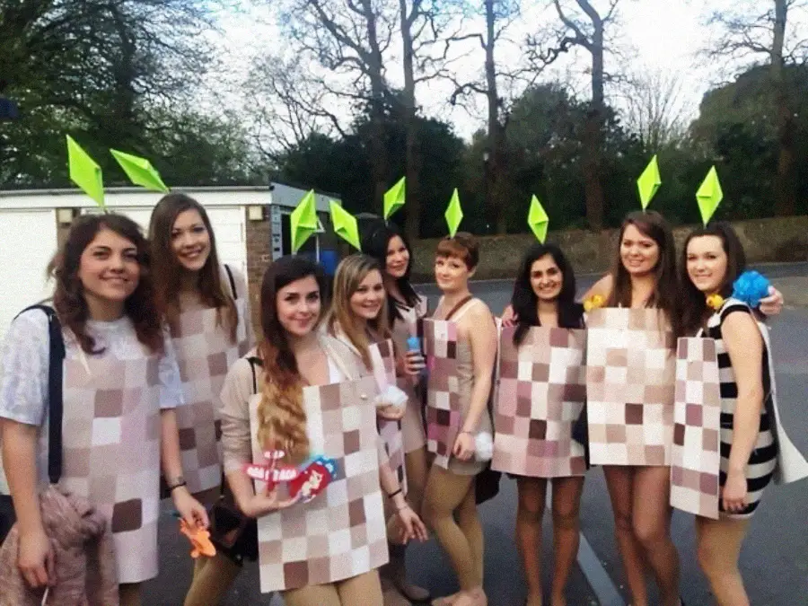 A group of people dressed as Sims characters, wearing pixelated outfits and green plumbob headpieces, stand together smiling outside on a paved area with trees in the background.