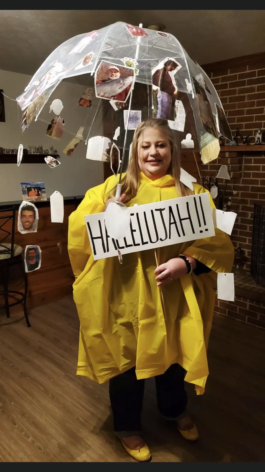 A woman in a yellow rain poncho holds a large sign reading "Hallelujah!!" She stands under a clear umbrella decorated with cut-out photos and tags, inside a room with a brick fireplace and wood panel walls.
