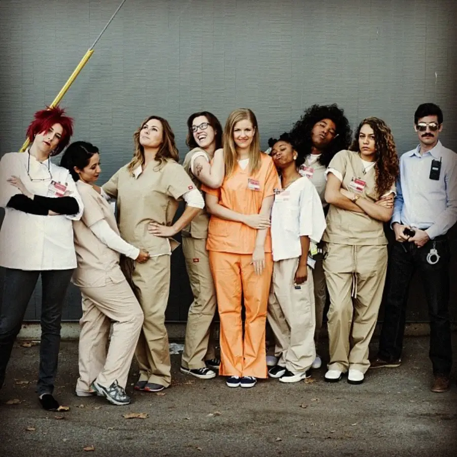Eight people dressed as characters from "Orange Is the New Black" pose together, with most wearing beige prison uniforms, one in orange, one in a lab coat, and one dressed as a guard with a mustache and handcuffs.