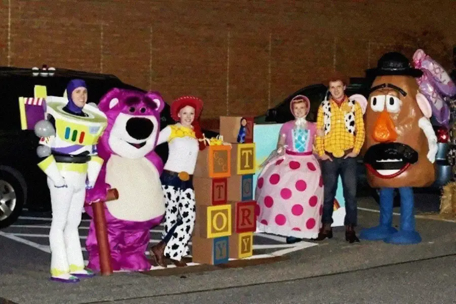 A group of people dressed as Toy Story characters—Buzz Lightyear, Lotso, Jessie, Bo Peep, Woody, and Mr. Potato Head—stand outdoors behind large toy blocks that spell "TOY STORY.