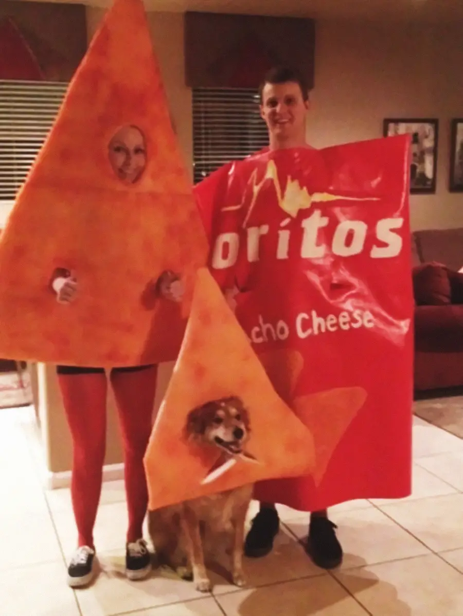 Two people in a living room wear Doritos-themed costumes—one as a nacho cheese Doritos bag, the other as a chip. A dog between them wears a matching chip costume. All appear to be smiling and posing for a photo.