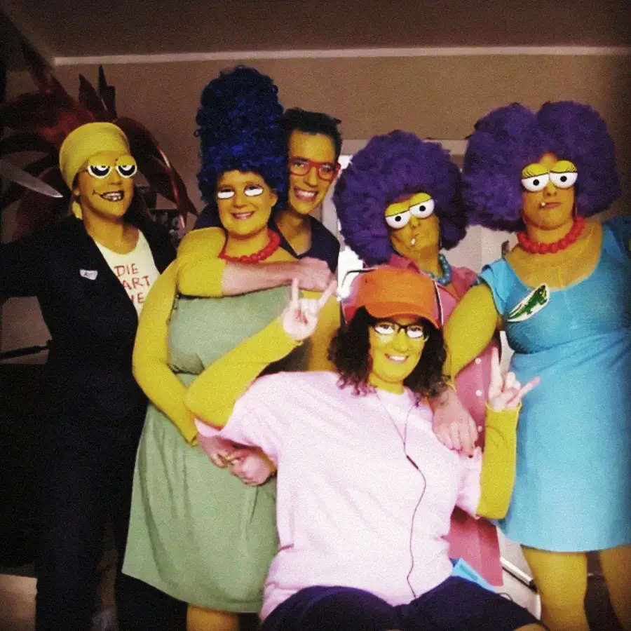 Six people wearing costumes and face paint to look like characters from The Simpsons. They have yellow skin, colorful wigs, and cartoonish glasses, recreating characters like Marge, Patty, and Selma. They pose together in a living room.