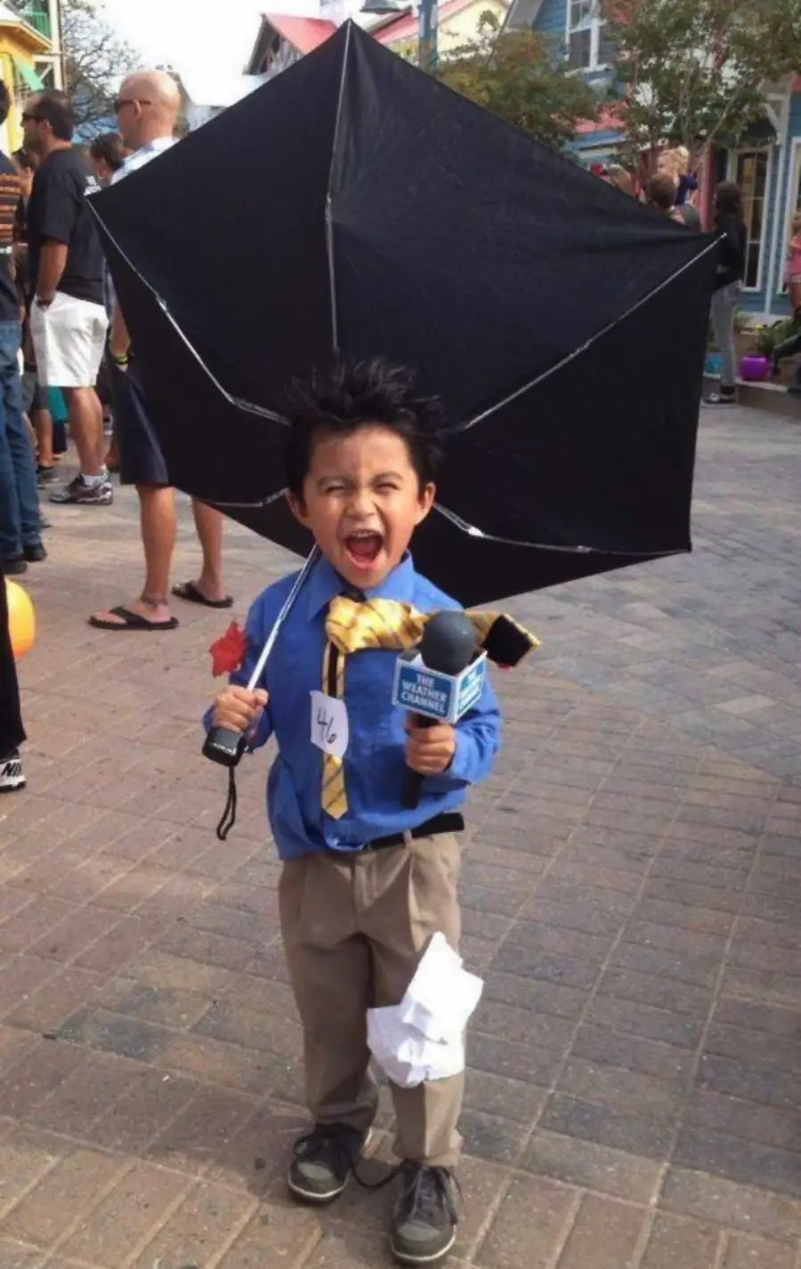 A young child in a blue shirt and yellow tie holds a broken umbrella and a microphone, pretending to be a news reporter in a storm. Paper and a name tag are attached to the outfit, and people are visible in the background.
