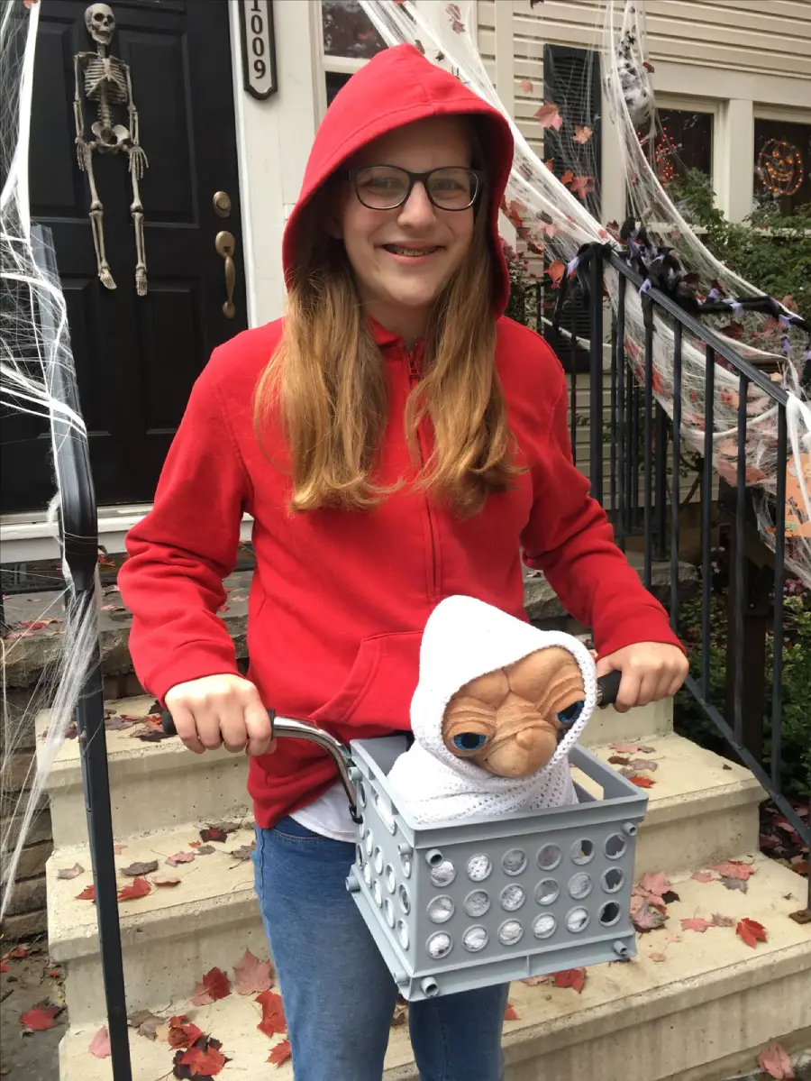 A person in a red hoodie stands on a stoop decorated for Halloween, holding a basket with a stuffed E.T. toy inside, recreating a scene from the movie "E.T. the Extra-Terrestrial.
