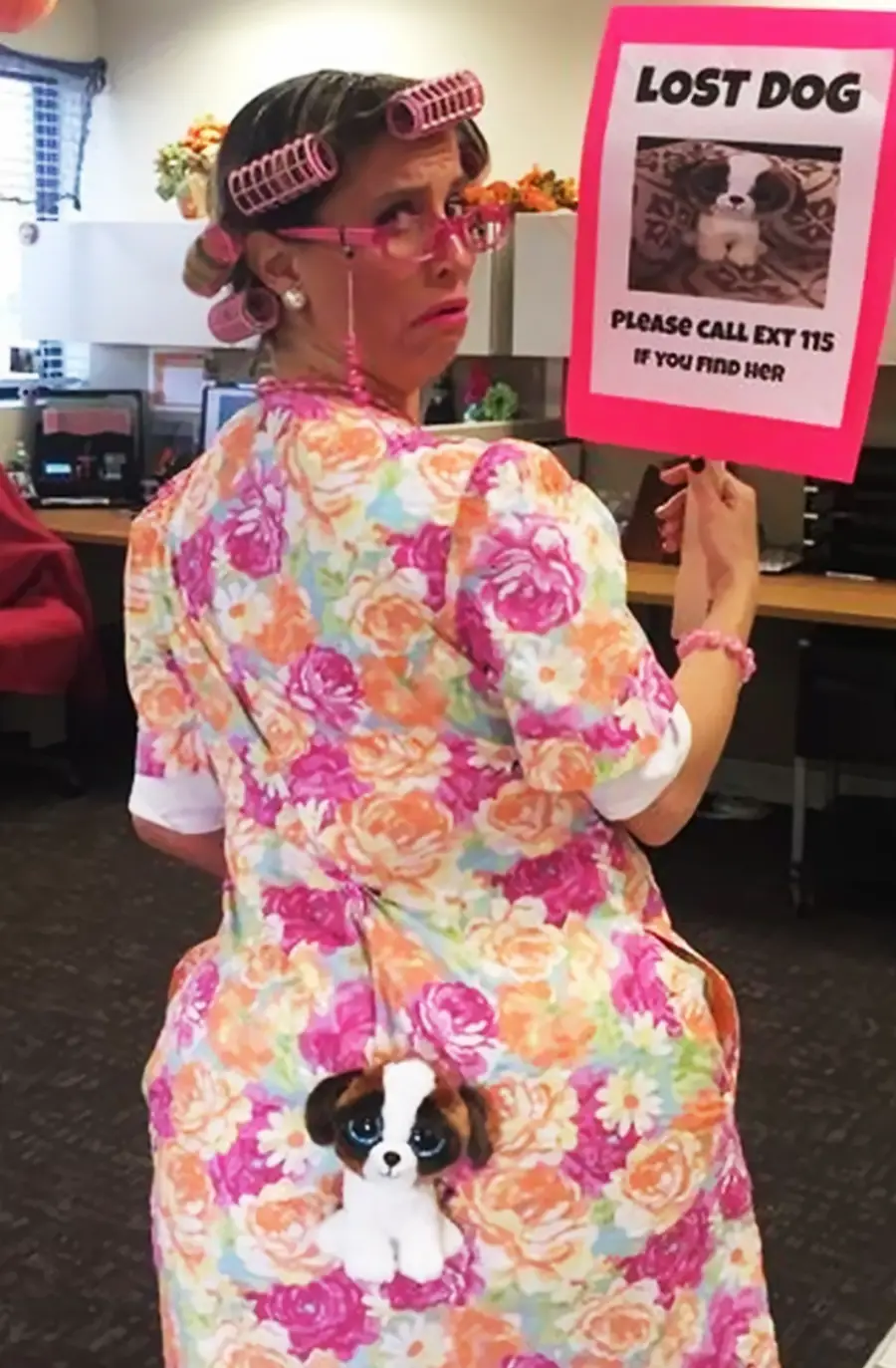 A woman in pink hair rollers and floral dress holds a "Lost Dog" sign. A small stuffed dog toy is attached to the back of her dress. She stands in an office, looking back over her shoulder with a surprised expression.