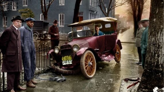 A vintage colorized photo shows several men standing near a damaged old car with a bent wheel on a wet city street, with tools and a detached tire lying on the sidewalk.