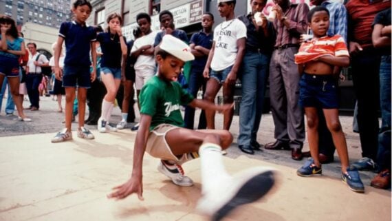 A young boy breakdances on a cardboard mat outdoors, surrounded by a group of children and adults watching intently. The scene has an urban setting with buildings in the background.