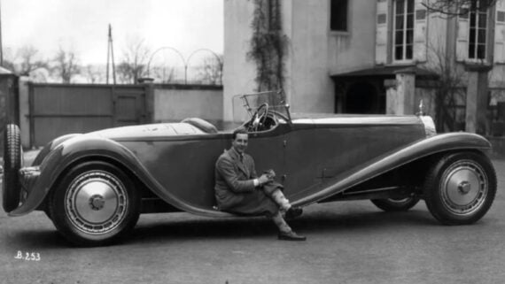 A vintage black-and-white photo of a person sitting on the side running board of a long, classic convertible car parked in front of a building with tall windows. The person is smiling and wearing a coat and boots.