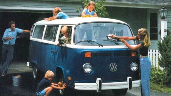A family washes a blue and white Volkswagen van in a driveway. Children clean the windows, wheels, and roof while a man and woman help nearby. A house and greenery are visible in the background.
