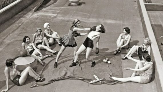 Two women in vintage swimwear and boxing gloves playfully spar while others sit around them on a rooftop, smiling and watching. Various props, including balls and ropes, are scattered on the ground.
