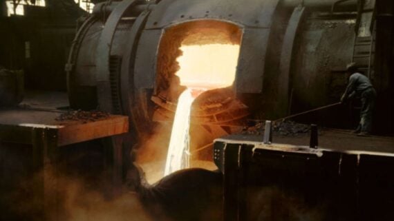 A worker stands near a large industrial furnace pouring a stream of glowing molten metal into a container in a factory setting.