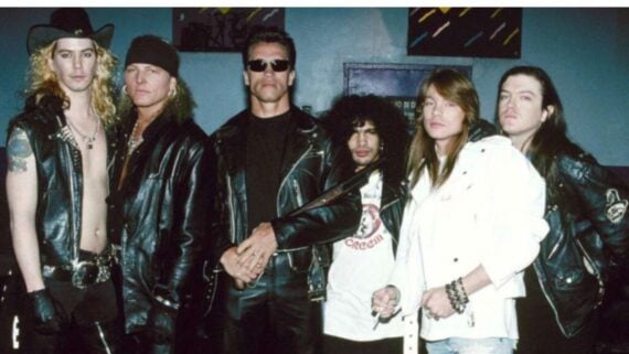 Six men stand side by side wearing mostly black leather jackets. One man in the center wears sunglasses and a black shirt. The group has long hair and rock band style, posing against a blue background.