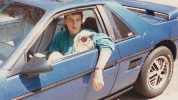 A young man sits in the driver&rsquo;s seat of a blue sports car with the window down. A small dog wearing sunglasses rests its paws on the window beside him.