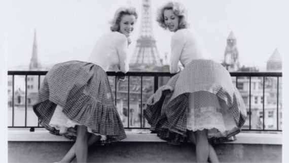 Two women in mid-20th-century outfits, with full plaid skirts and white blouses, lean over a balcony railing, smiling, with the Eiffel Tower and Paris skyline visible in the background.