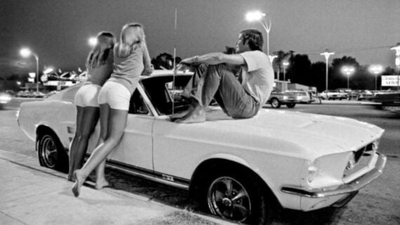 Two young women in shorts stand by a classic white car as a man sits barefoot on the hood. The scene is set at night on a street illuminated by streetlights, with other cars and buildings in the background.