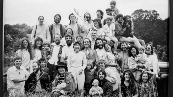 A large group of people, including adults and children, pose closely together outdoors, smiling and laughing. The image is black and white, and they appear to be in a rural, grassy area with trees in the background.
