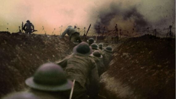 Soldiers wearing helmets move through a muddy trench during battle, with barbed wire ahead and smoke rising from explosions in the distance, suggesting an intense wartime scene.