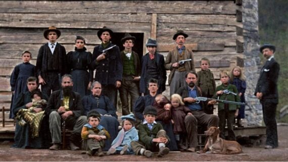 A group of men, women, and children, some holding rifles, pose in front of a rustic wooden cabin. They wear old-fashioned clothing; a dog lies at their feet. The background is forested and mountainous.