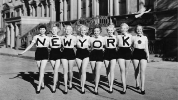 Eight women dressed in matching outfits stand in a row on a city street, each holding a large letter to collectively spell "NEW YORK" with a dot at the end. Classic black-and-white photo, likely from the mid-20th century.