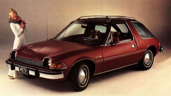 A woman in a white outfit stands next to a maroon 1970s AMC Pacer car in a studio setting with a neutral background.