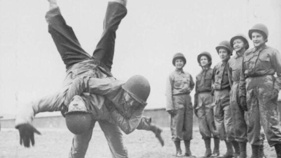 A group of women in military uniforms and helmets watch as one woman flips another during a martial arts or self-defense demonstration outdoors.